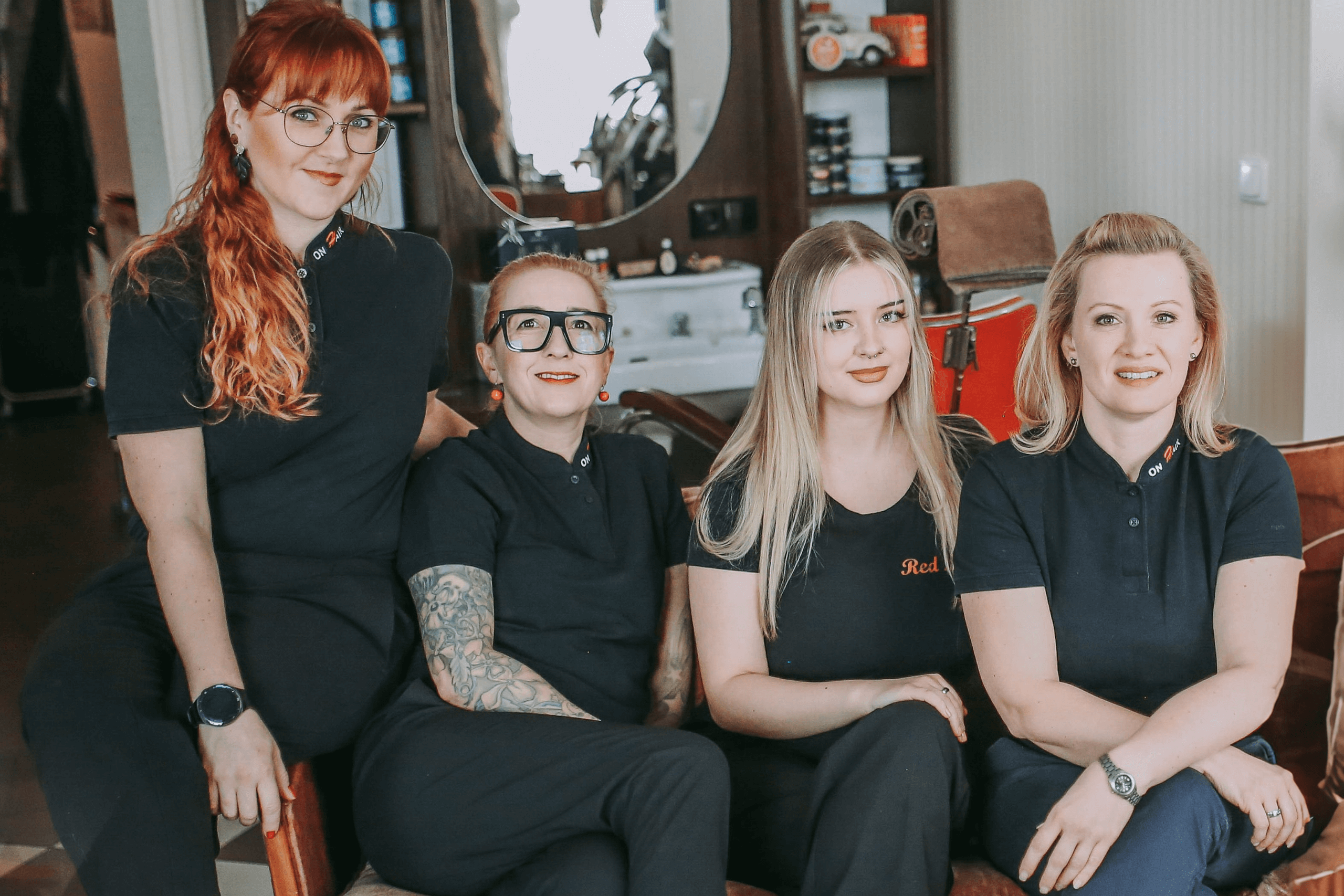 Vier Frauen des OnHair Teams sitzen gemeinsam in einem Friseursalon auf einem Ledersessel. Sie tragen einheitlich dunkle Kleidung mit dem Markenlogo und lächeln in die Kamera. Im Hintergrund sind ein Spiegel, ein Waschbecken und Pflegeprodukte sichtbar.